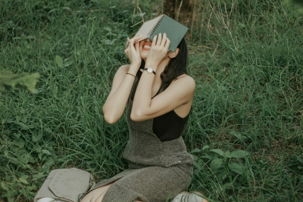 Woman in Black Tank Top and Gray Pants Sitting on Grassfield Reading Book